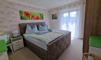 A cozy bedroom with a large double bed and bright colors. Pictures with floral motifs hang on the wall, and large windows let in plenty of light.