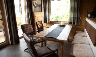 A cozy dining area in a wooden house with a wooden table and several chairs. The windows offer a beautiful view of the greenery.
