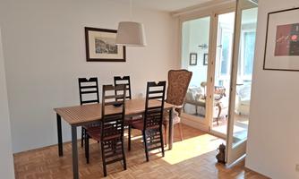 A bright dining area with a wooden table and six chairs. Large windows ensure plenty of natural light and connect the space with an adjacent living area.