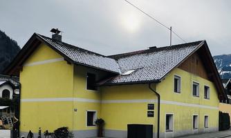 A yellow house with a snow-covered roof on a quiet street. In the background, mountains and a cloudy sky can be seen.