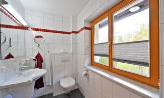 A modern bathroom with white tiles and a large window. It features a sink, toilet, and elegant towels in red.