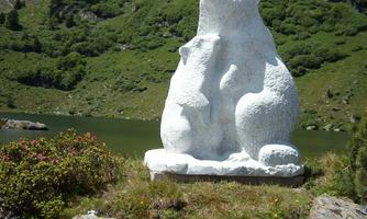 A large, white bear statue stands on a meadow by the water. In the background, green hills and blue sky are visible.