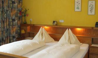 A cozy bedroom with a double bed and two large, white pillows. The walls are painted in a warm yellow tone, and there are decorative elements such as pictures and curtains.