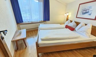 A cozy bedroom with two single beds and wooden furniture. The room has large windows with curtains and a wooden table.