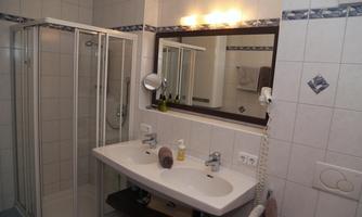 A modern bathroom with a shower cabin and a double sink. The mirror reflects the bright, tiled wall.