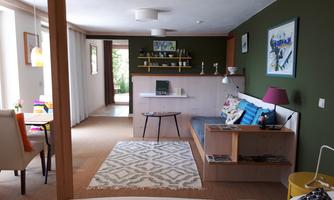 A modern furnished living and dining room with a cozy sofa, a table, and colorful chairs. The walls are painted in a dark green shade, and there is plenty of daylight.