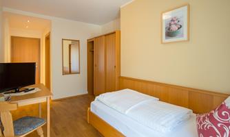 A bright room with a single bed, a table, and a television. On the wall hangs a picture with floral motifs.