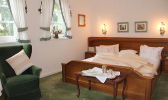 A cozy bedroom with a large wooden bed and fresh white bedding. In the corner stands a green armchair, and the windows are adorned with elegant curtains.