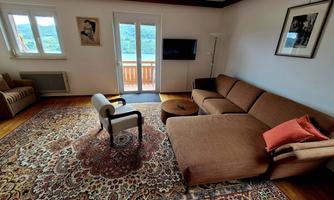 A cozy living room with a large sofa and a round table. There is a rug and windows that lead to a balcony.