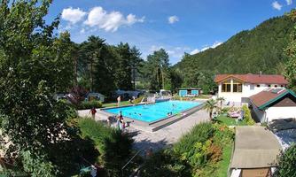 A swimming pool surrounded by trees and mountains. People enjoy the sun and the water.