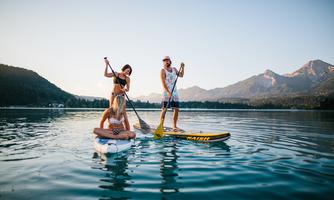 Three people are paddling on a calm lake in front of a picturesque mountain backdrop. It is a sunny day and the atmosphere is relaxed.