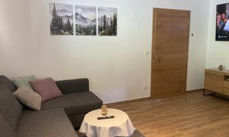 A cozy living room with a gray sofa and a small coffee table. Landscape pictures hang on the wall and a wooden door leads outside.