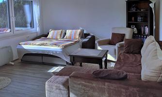 A cozy living room with a sofa bed and various seating options. The room has large windows that let in plenty of light.