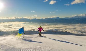 Two skiers are skiing on a snow-covered slope. In the background, the mountains stretch out and the sky is clear and sunny.