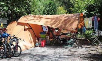 A large tent in a green camping area, surrounded by trees. Next to the tent are bicycles and a clothesline with clothes.