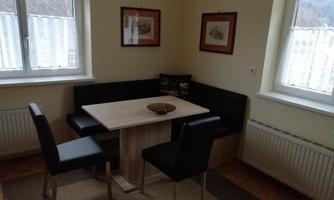 A small dining area with a table and chairs. Pictures hang on the walls, and there are large windows that let in light.