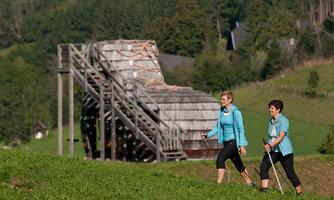 Two women are hiking on a green hill. In the background, a large wooden structure is visible.