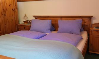 A cozy bedroom with a wooden bed and purple pillows. Next to the bed stands a wooden wardrobe and a nightstand.