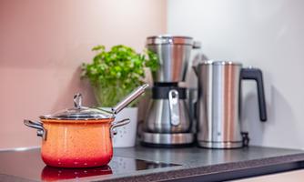 A modern kitchen with a colorful pot on the stove and a coffee machine in the background. Fresh herbs complement the decor.