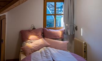 A cozy bedroom with a bed decorated with pink pillows and blankets. A window overlooking the surroundings provides natural light.