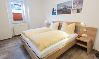 A modern bedroom with a double bed and large, comfortable pillows. Landscape pictures hang on the wall, and there is a nightstand with a lamp.