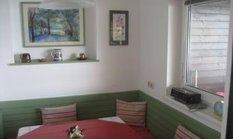 A cozy dining area with a green bench and a round table. A picture hangs on the wall, and there is a window with a view outside.