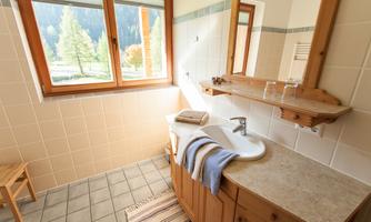 A bright bathroom with a large window and wooden shelves. The sink is decorated with towels and a small rug.