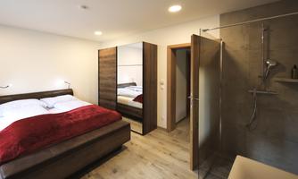 A modern bedroom with a large, comfortable bed and warm, red blankets. On the right side there is an adjacent shower and a wooden wardrobe.