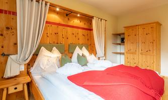 A cozy bedroom with a double bed, wooden furniture, and a warm red blanket. The walls are clad in wooden panels and there are light curtains.