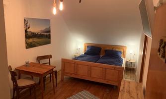 A cozy room with a double bed, wooden furniture, and a small seating area. A picture of a landscape hangs on the wall, and the floor is covered with a carpet.