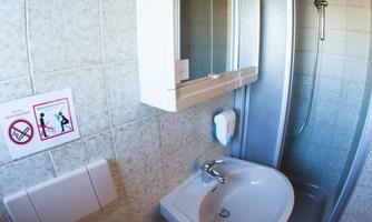 A small bathroom with shower, sink, and toilet. The walls are light tiled, and there is a wood ceiling paneling.