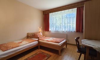 A simple room with two single beds and a table. Large windows bring in daylight and the floor is made of wood.
