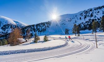 A snow-covered landscape with shimmering sun. Two people are skiing on a winding track.
