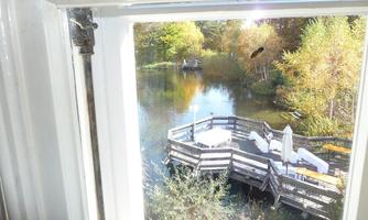 A view from a window onto a calm lake with a wooden terrace. In the background, trees with colorful leaves can be seen.