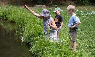 An adult is fishing by the shore of a pond. Two children are watching eagerly.