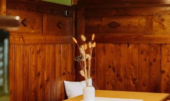 A cozy wooden dining table with chairs in a warm room. On the table is a vase with dried flowers and a tablecloth.