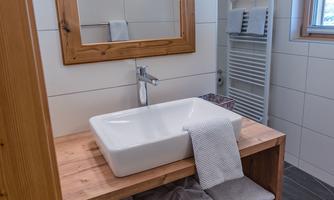 A modern bathroom with a wooden vanity and a rectangular sink. Clean towels are neatly arranged on the shelf, and there is plenty of daylight.