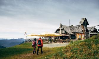 A pair of hikers is walking to a mountain lodge with a terrace. In the background, the mountains and a cloudy sky are visible.