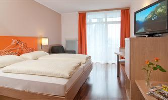 A cozy hotel room with two single beds and modern furniture. There are large windows with curtains and a television on the wall.