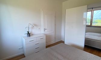 A modern bedroom with a large bed and a white wardrobe. The room has plenty of natural light and a calm atmosphere.