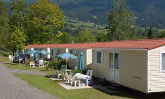 A campsite with several mobile homes in a green landscape. In the foreground, there are tables and chairs under colorful sun umbrellas.