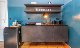 A modern kitchen with a black kitchen base cabinet and an open shelf. The walls are painted blue and the floor is made of wood.