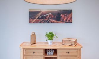 A cozy room with a wooden table, on which a plant in a pot is standing. A landscape painting with sun rays and autumn colors hangs on the wall.
