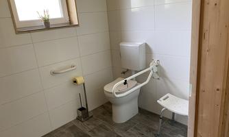 The image shows a modern bathroom with an accessible toilet. It has bright tiles, a window, and a seating area.