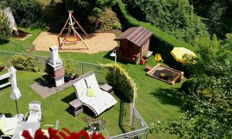 A beautiful garden with a playground, swing, and a cozy little house. There are loungers, a barbecue area, and sunny spots for relaxing.
