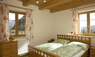 A cozy bedroom with wooden furniture and a large window. The bedding has a cheerful, green pattern and the walls are painted in a light color.
