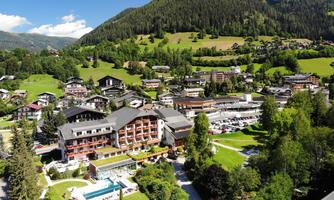 A picturesque village in the mountains with a modern hotel and a pool. The surroundings are green fields with trees and a beautiful landscape.