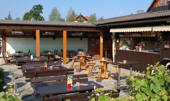 A cozy beer garden with wooden chairs and tables under a blue sky. In the background, a bar with various drinks is visible.