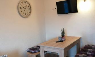 A cozy room with a wooden table and benches. A clock is hanging on the wall, and a television is installed.