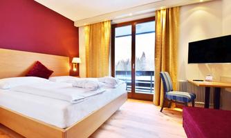 A modern hotel room with a large bed and a cozy armchair. Large windows let in plenty of light and provide access to a balcony.